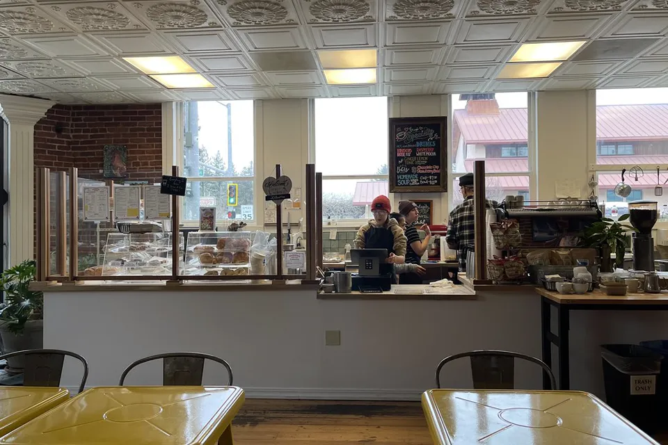Cafe ordering counter seen past yellow metal tables, with pastry display cases, a chalkboard menu and baristas working by bright windows.