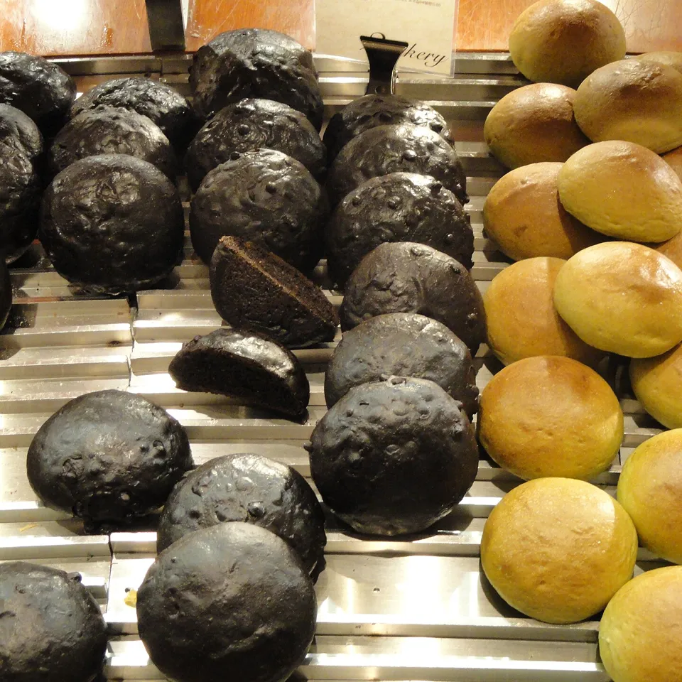 Rows of dark chocolate-glazed round buns, one cut open, beside golden round buns on a bakery's steel display rack in Seoul.