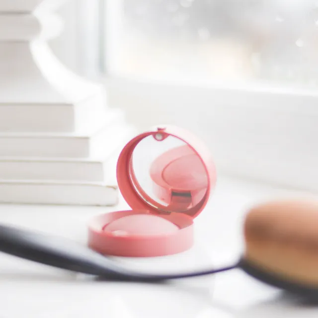 Open pink blush compact with a mirror on a white windowsill, oval makeup brush blurred in front, stacked books behind