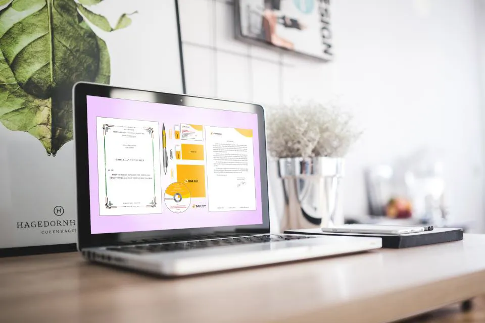 Laptop on a wooden desk showing stationery and branding design mockups, dried flowers in a silver pot, bright minimal room
